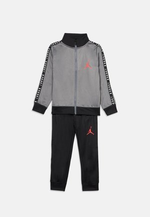 Pilkas sportinis kostiumas su juodais akcentais, su užtrauktuku užsegama striuke ir derančiomis kelnėmis. Raudonas logotipas ant striukės ir kelnių. Lygus audinys, sportinis dizainas.