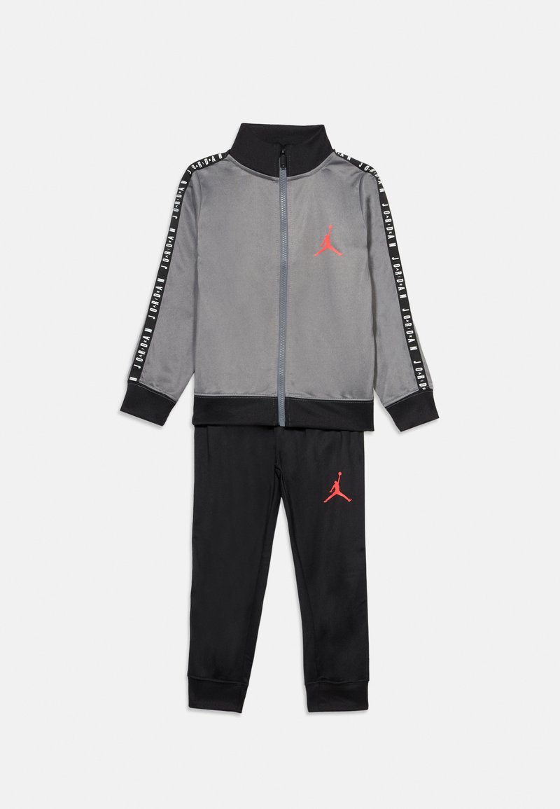 Γκρι φόρμα με μαύρες λεπτομέρειες, που περιλαμβάνει ζακέτα με φερμουάρ και παντελόνι που ταιριάζει. Κόκκινο λογότυπο στη ζακέτα και το παντελόνι. Λείο ύφασμα, αθλητικός σχεδιασμός.