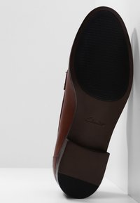 Chaussure en cuir marron avec une semelle extérieure en caoutchouc noir texturé. Présente un bout arrondi et un petit talon, mettant en valeur un design minimaliste et un savoir-faire artisanal.