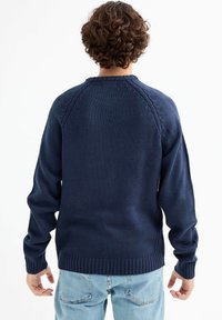 Suéter navy con puños y dobladillo acanalados. Cuenta con mangas raglán y un patrón de tejido texturizado. Llevado sobre jeans azul claro. Vista trasera.