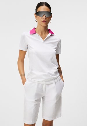 Weißes Poloshirt mit rosa Kragen, kombiniert mit weißen Shorts. Das Material wirkt glatt und leicht, geeignet für legere Anlässe.