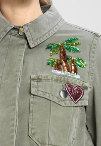 Oliivihreä paita, jossa on paljettiompelulla koristeltu palmu puhelinjärjestelmän taskussa ja sydämen muotoinen merkki. Napitus ja kaulus.