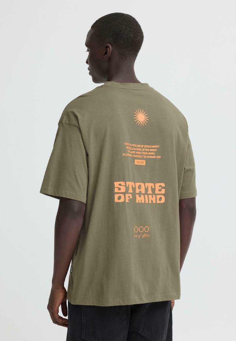 Moški, oblečen v jopico olivno zelene barve z oranžnim napisom in sončnim grafikonom, viden od zadaj, na preprostem svetlem ozadju.