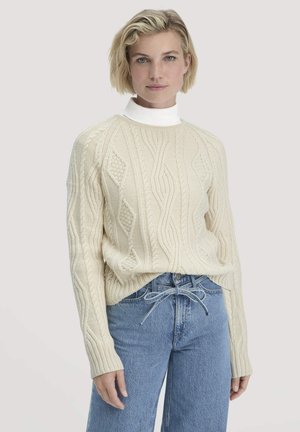 Crème kabelgebreide trui met een hoge nek, met textuurpatronen. Gekoppeld aan lichtblauwe jeans en een zichtbare tailleband.