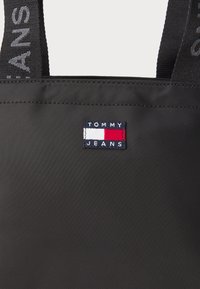 Schwarze Tragetasche mit strukturiertem Oberflächen, versehen mit einem rechteckigen gestickten Logo in Rot, Weiß und Blau mit der Aufschrift "TOMMY JEANS."