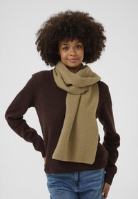 Strikket tørklæde i beige med ribbet tekstur, hængende om halsen. Båret over en mørkebrun sweater og blå jeans.