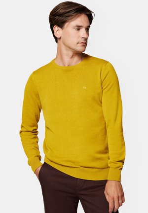 Żółty sweter z dzianiny, wykonany z miękkiego materiału, z okrągłym dekoltem, prążkowanymi mankietami i małym logo na klatce piersiowej. Noszony z brązowymi spodniami.