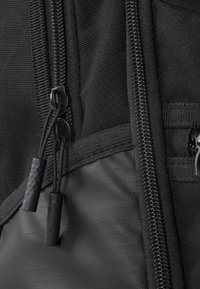 Fekete szövet hátizsák részlet, amely cipzárakat mutat textúrált húzófülekkel, anyagtípusok közötti kontrasztos textúrákkal és elegáns kialakítással.