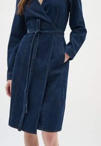 Denimklänning i mörkblått med långa ärmar, en midja med bälte och sidofickor. Har synlig sömnad och en flikad krage.