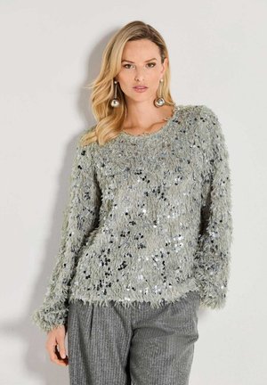 Woman with blonde hair wearing a grey fuzzy sweater with sequins and large silver dangling earrings, standing against a plain light background.