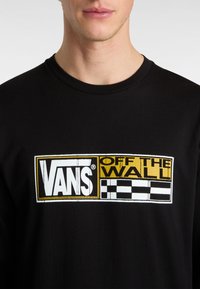 Schwarzes Langarm-T-Shirt mit einem weißen und gelben Vans-Logo und Schachbrettakzenten auf der Vorderseite. Hergestellt aus weichem Baumwollmaterial.