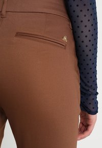 Hnědé kalhoty ve slim fit střihu, s jednou zadní kapsou a malým zlatým logem. Částečně viditelný texturovaný top s dlouhým rukávem v námořnické barvě s puntíky.