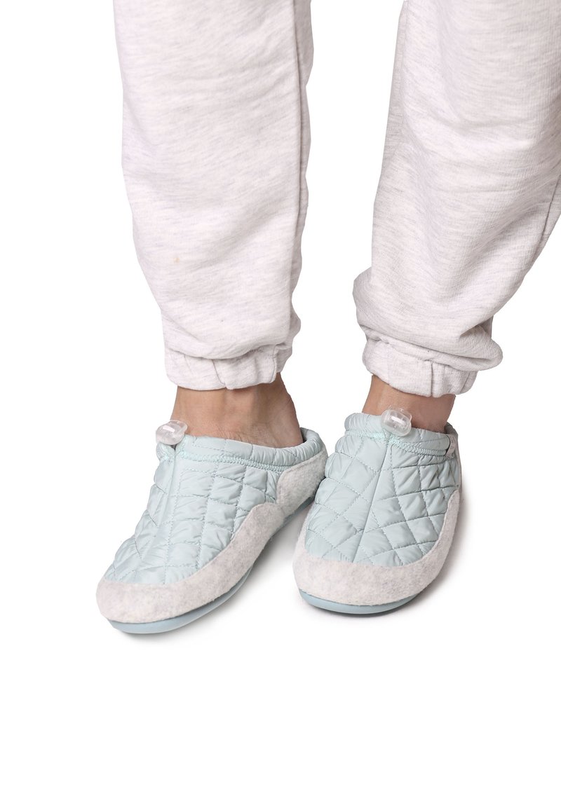 Pantofole di tessuto trapuntato blu chiaro con accenti in suede grigio e chiusura a bottone trasparente, abbinate a pantaloni jogger grigi.
