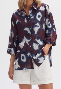 Chemise transparente à boutons avec un motif abstrait maroon, bleu et blanc, manches longues avec poignets, et coupe ample. Associée à un short blanc.