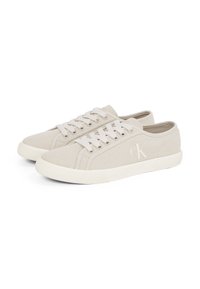 Lichtbeige lage canvas sneakers met witte veters, rubberen zolen en het "cK" logo aan de zijkant, naast elkaar geplaatst.