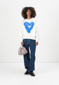 Felpa bianca con grafica a cuore blu e testo nero, abbinata a jeans di denim blu larghi e una piccola borsa lavanda. Scarpe nere completano il look.