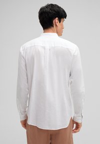 Camicia bianca a maniche lunghe in un tessuto leggero, dotata di colletto, polsini con bottone e un orlo leggermente curvo, mostrata da dietro.