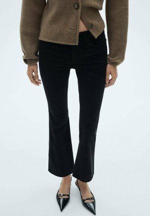 Person trägt schwarze, gekürzte Hose, braunen, geknöpften Pullover und schwarze spitze Schuhe mit Knöchelriemen, stehend vor einfarbig hellem Hintergrund.