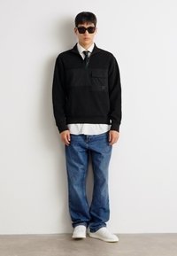 Pullover nero con zip e pannello testurizzato, indossato sopra una camicia bianca. Abbinato a jeans blue denim e sneaker bianche. Occhiali da sole neri molto grandi.