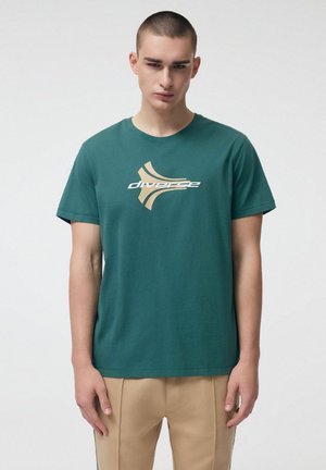 Młody mężczyzna w turkusowej koszulce z beżowo-białym abstrakcyjnym logo i beżowych spodniach, stojący na jednolitym jasnym tle.