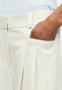 Χέρι με σκούρο δέρμα που ακουμπά μέσα στην μπροστινή τσέπη μπεζ παντελονιού με λεπτές ρίγες και θηλιές για ζώνη.