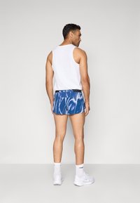 Männer-weißes Tanktop mit blauen gemusterten Shorts. Die Shorts haben einen schwarzen Bund und eine glänzende Stoffoberfläche. Weiße Sportsocken und Schuhe.