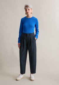 Niebieski, prążkowany top z długim rękawem w połączeniu z czarnymi, wysokim stanem spodniami i białymi trampkami. Modelka nosi kratkowaną czapkę. Proste tło.