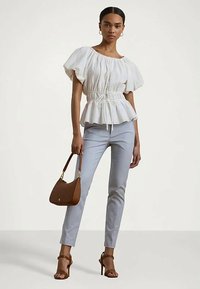 Γυναίκα που φοράει λευκή μπλούζα με φουσκωτά μανίκια και σύστημα ρύθμισης στη μέση, ανοιχτού γκρίζου κομψά παντελόνια, καφέ ψηλοτάκουνα σανδάλια, κρατώντας καφέ τσάντα.