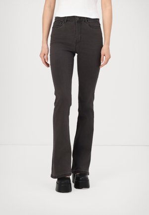 Žena oblečená v černých rozšířených džínách s vysokým pasem, doplněných černými platformovými botami a bílým topem, stojící před jednobarevným světlým pozadím.