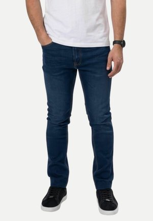 Homme portant un jean slim bleu foncé, des baskets noires à semelles blanches et une chemise blanche, debout avec une main dans la poche.