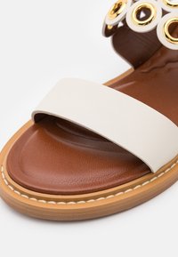 Sandale en cuir marron avec une bride blanche au niveau des orteils et des éléments circulaires décoratifs blancs et dorés près de la cheville.