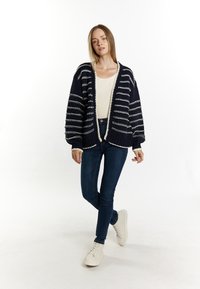 Cardigan a righe navy con accenti crema, apertura frontale, tessuto testurizzato e spalle scese. Abbinato a jeans slim e sneaker chiare.
