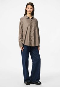 Rjava in bež prugasta srajca s kratkimi rokavi in ovratnikom, v kombinaciji s širokimi modrimi kavbojkami, ki ponujajo sproščen kroj in žep na prsih.