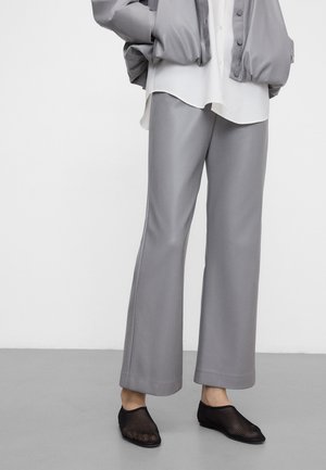 Osoba ubrana w luźne szare spodnie, białą koszulę, szarą kurtkę i czarne siateczkowe buty wsuwane, stojąca na szarej podłodze.