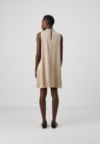 Beige mekko ilman hihoja, jossa on avainaukko selkäosassa, sileä satiinimateriaali, hillitty kiilto ja minimalistinen muotoilu, joka ulottuu polven ylle.