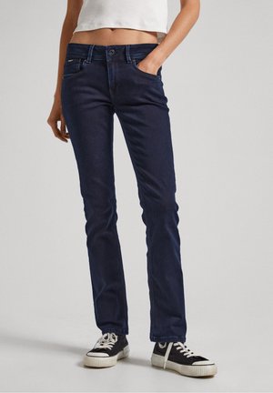 Kobieta ubrana w ciemnoniebieskie jeansy o prostych nogawkach, biały krótki top oraz czarne trampki z białymi sznurówkami, stojąca z jedną ręką w kieszeni.