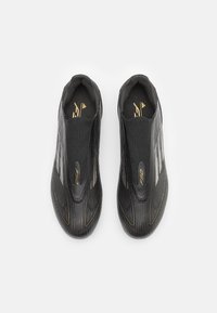 Zapatillas deportivas negras con un diseño elegante, que presentan rayas verticales, una textura suave y detalles en dorado. Estilo sin cordones con una marca minimalista.