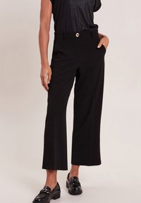 Zwarte wijde broeken met een gladde textuur, voorzien van een sluiting met een knoop aan de voorkant en zijzakken, gestyled met een losse zwarte top en zwarte loafers.