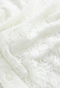 Balts mežģīņu audums ar sarežģītām ziedu rakstiem, tekstūru sajaukumu un gludu apdari ar redzamu pogas detaļu.