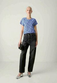 Mulher a usar uma blusa curta de mangas azuis com padrão floral, jeans pretos de cintura alta, tacões prateados em bloco, brincos em argola e a segurar uma pequena mala preta.