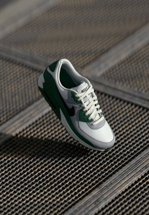 Białe i zielone sneakersy Nike Air Max z teksturowaną skórzaną cholewką, czarnymi akcentami i charakterystycznym białym systemem sznurowania. Ułożone nad siateczkową powierzchnią.