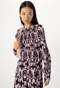 Langärmliges Kleid mit einem rosa und schwarzen Blumenmuster, plissiertem Design und rundem Ausschnitt, gehalten mit einem hellbeigen Träger.