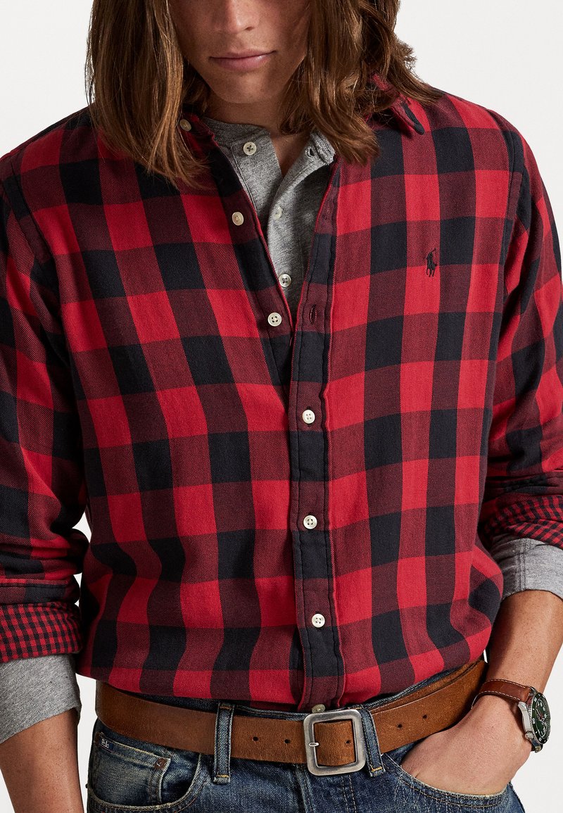 Rød og sort ternet flannel skjorte med knaplukning foran; grå henley nedenunder; brun bælte; afslappet denimlook; lange ærmer med kontrasterende manchetter.