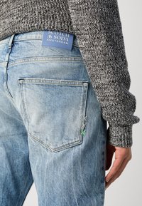 Un jean en denim bleu clair associé à un pull gris texturé. Les caractéristiques incluent un détail de poche cousue et une étiquette de marque bleue sur la ceinture.