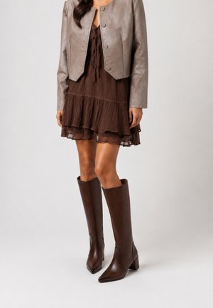 Frau trägt braunes, gestuftes Kleid, kurze metallische Jacke und kniehohe braune Lederstiefel, steht vor einfachem Hintergrund.