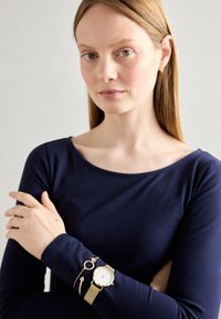 Kuldse tooni kell valge dialiga beežist võrgust rihmaga, koos kuldse banglese käevõruga. Pikkade varrukatega tumeintonine särk, millel on istuv disain.