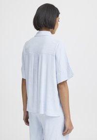 Femme debout, de dos, portant une chemise à manches courtes ample bleu clair et un pantalon assorti, sur un fond clair uni.