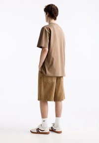 Camiseta de algodón marrón, ajuste oversize, combinada con pantalones cortos de mezclilla beige. Zapatillas blancas con acentos en color burdeos y calcetines blancos. Diseño simple, sin patrones.