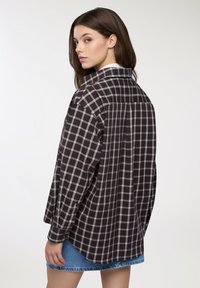 Camicia oversize a scacchi in bianco e nero con colletto e polsini abbottonati, con un lungo retro e vestibilità rilassata. Indossata con una gonna in denim.