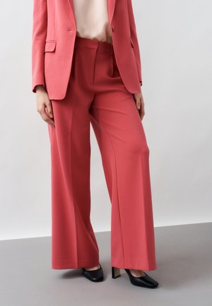 Donna che indossa pantaloni larghi color corallo, blazer coordinato, top beige e scarpe con tacco a blocco nere, in piedi davanti a uno sfondo neutro.